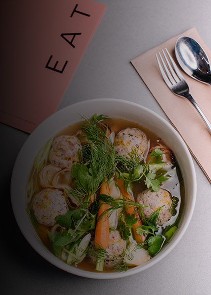 Bowl of hearty noodle soup with meatballs, fresh herbs, and vegetables, placed on a gray surface next to silverware and a napkin