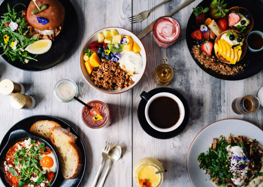 Overhead view of a colorful brunch spread featuring fresh fruit bowls, a burger with greens, toast with eggs, coffee, and smoothies, arranged on a rustic wooden table
